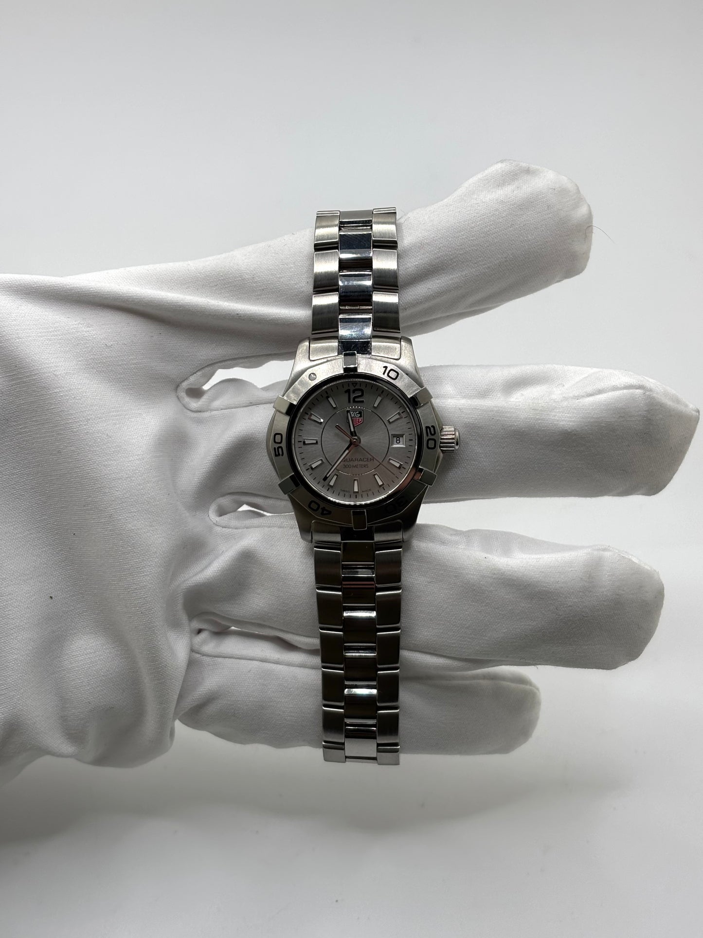 Montre femme TAG Heuer Aquaracer Lady référence WAF1412 en acier avec boîtier 27 mm et bracelet acier. Cadran argent avec chiffres arabes, mouvement quartz précis et étanchéité 30 ATM. Montre de luxe sportive et élégante en très bon état, révisée en 2026, idéale pour un usage quotidien ou une tenue habillée. Détail du boîtier acier poli, montre horlogère suisse féminine, intemporelle et raffinée.