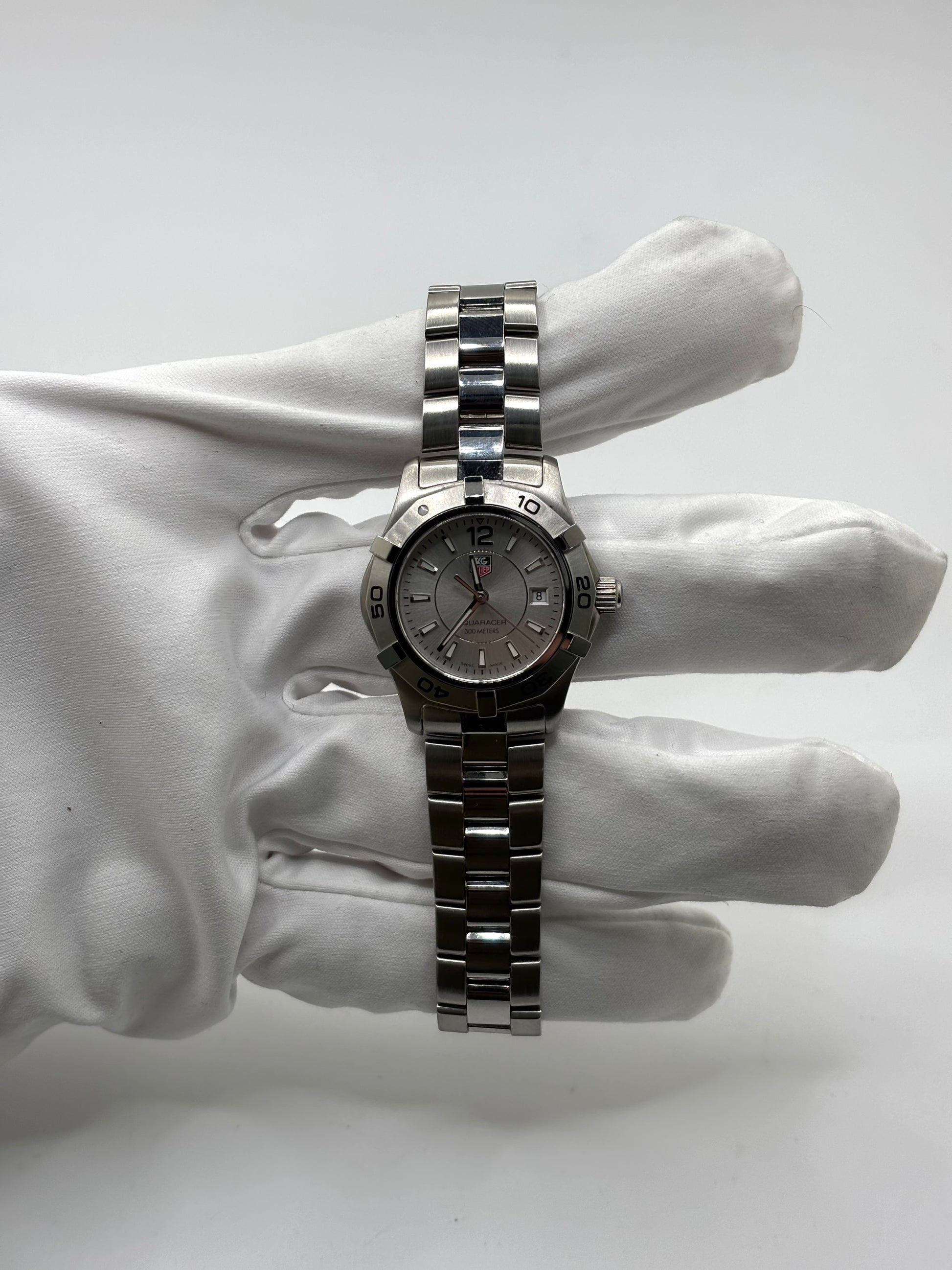 Montre femme TAG Heuer Aquaracer Lady référence WAF1412 en acier avec boîtier 27 mm et bracelet acier. Cadran argent avec chiffres arabes, mouvement quartz précis et étanchéité 30 ATM. Montre de luxe sportive et élégante en très bon état, révisée en 2026, idéale pour un usage quotidien ou une tenue habillée. Détail du boîtier acier poli, montre horlogère suisse féminine, intemporelle et raffinée.