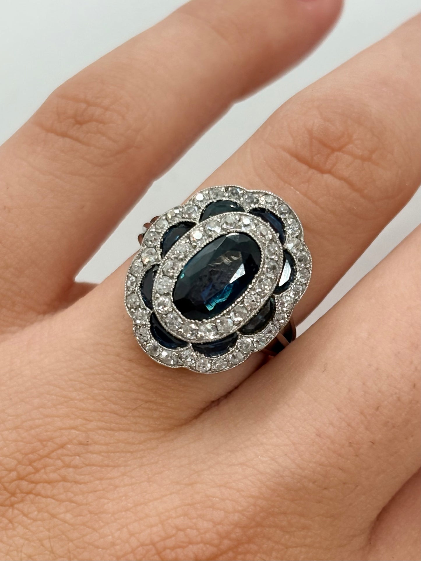 Bague ancienne en or blanc 18K (750 millièmes), sertie d’un saphir naturel central ovale d’environ 2 carats et de petits saphirs naturels en demi-lune. Des diamants naturels de taille ancienne complètent cette pièce de joaillerie pour un poids total d’environ 0,25 carat. Cette bague vintage en or blanc séduit par son élégance intemporelle, ses pierres précieuses et son charme raffiné.