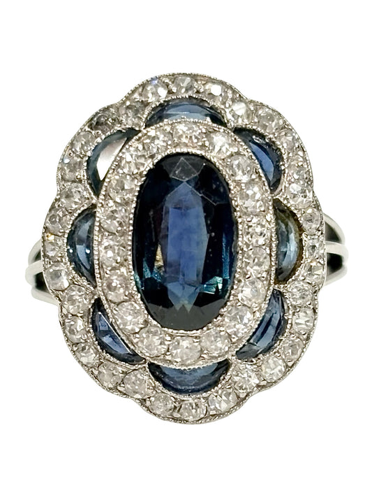 Bague ancienne en or blanc 18K (750 millièmes), sertie d’un saphir naturel central ovale d’environ 2 carats et de petits saphirs naturels en demi-lune. Des diamants naturels de taille ancienne complètent cette pièce de joaillerie pour un poids total d’environ 0,25 carat. Cette bague vintage en or blanc séduit par son élégance intemporelle, ses pierres précieuses et son charme raffiné.
