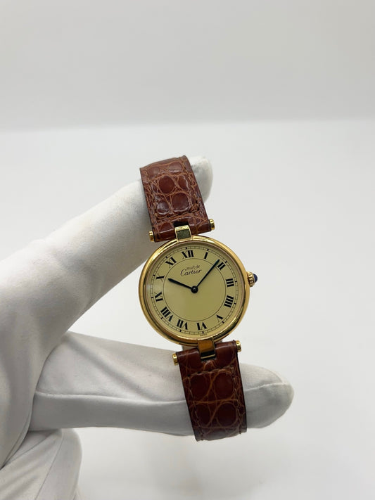 Montre Cartier Must Vendôme réf. 590003 en argent, cadran doré à chiffres romains, boîtier 30 mm avec verre saphir. Mouvement quartz fiable, bracelet cuir noir avec boucle ardillon. Modèle vintage de 1990 en très bon état, avec légères micro-rayures peu visibles. Livrée avec coffret et papiers d’origine, révisée en 2026.