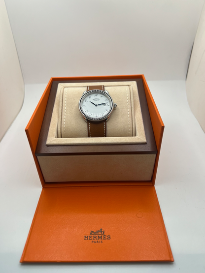 Montre Hermès Arceau Grand Modèle AR5.710 de 2014 en acier 38 mm avec mouvement quartz, cadran blanc à chiffres arabes et bracelet cuir brun d’origine. Cette montre de luxe Hermès Full Set avec boîte et papiers possède un verre saphir, une boucle ardillon acier et le design emblématique Arceau inspiré de l’univers équestre. Montre vintage Hermès élégante, révisée en 2026 et garantie 1 an.