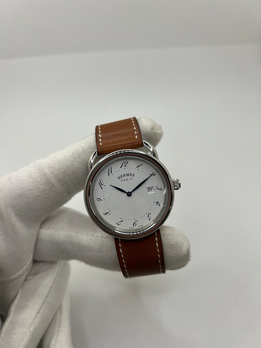 Montre Hermès Arceau Grand Modèle AR5.710 de 2014 en acier 38 mm avec mouvement quartz, cadran blanc à chiffres arabes et bracelet cuir brun d’origine. Cette montre de luxe Hermès Full Set avec boîte et papiers possède un verre saphir, une boucle ardillon acier et le design emblématique Arceau inspiré de l’univers équestre. Montre vintage Hermès élégante, révisée en 2026 et garantie 1 an.