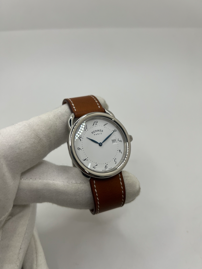 Montre Hermès Arceau Grand Modèle AR5.710 de 2014 en acier 38 mm avec mouvement quartz, cadran blanc à chiffres arabes et bracelet cuir brun d’origine. Cette montre de luxe Hermès Full Set avec boîte et papiers possède un verre saphir, une boucle ardillon acier et le design emblématique Arceau inspiré de l’univers équestre. Montre vintage Hermès élégante, révisée en 2026 et garantie 1 an.
