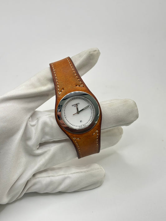 Montre Hermès Harnais HA3.410 en acier avec mouvement quartz, cadran blanc et bracelet en cuir brun. Modèle homme et unisexe au design élégant avec verre saphir et fonction date. Montre d’occasion en très bon état avec légères traces d’usure et micro-rayures discrètes. Format 30 mm idéal pour un style classique et raffiné.

