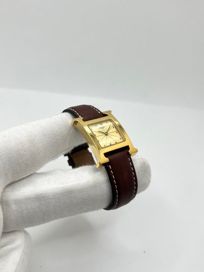 Montre Hermès Heure H HH1.201 pour femme, équipée d’un mouvement quartz et d’un boîtier en acier de 21 mm. Son cadran couleur or est protégé par un verre saphir et accompagné d’un bracelet brun en cuir de vache avec boucle ardillon en acier. Étanche à 3 ATM, cette montre Hermès Heure H se distingue par son célèbre boîtier en forme de H et son design élégant. En très bon état, elle présente de légères traces d’usure.
