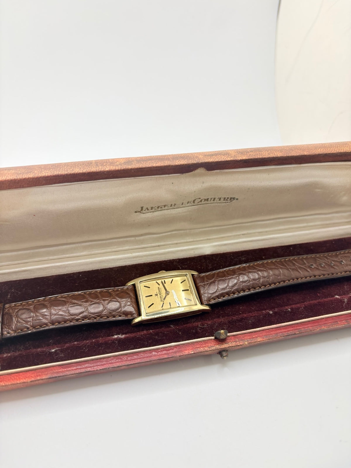 Montre Jaeger-LeCoultre Classic 9012 vintage à remontage manuel, datant approximativement de 1960. Son boîtier rectangulaire en or jaune de 37 × 23 mm est équipé d’un verre Plexiglas et d’un élégant cadran or à index bâtons. Cette montre Jaeger-LeCoultre est montée sur un bracelet brun en cuir de crocodile avec boucle ardillon en acier. En très bon état, elle présente de légères traces d’usure et est proposée avec son coffret d’origine.