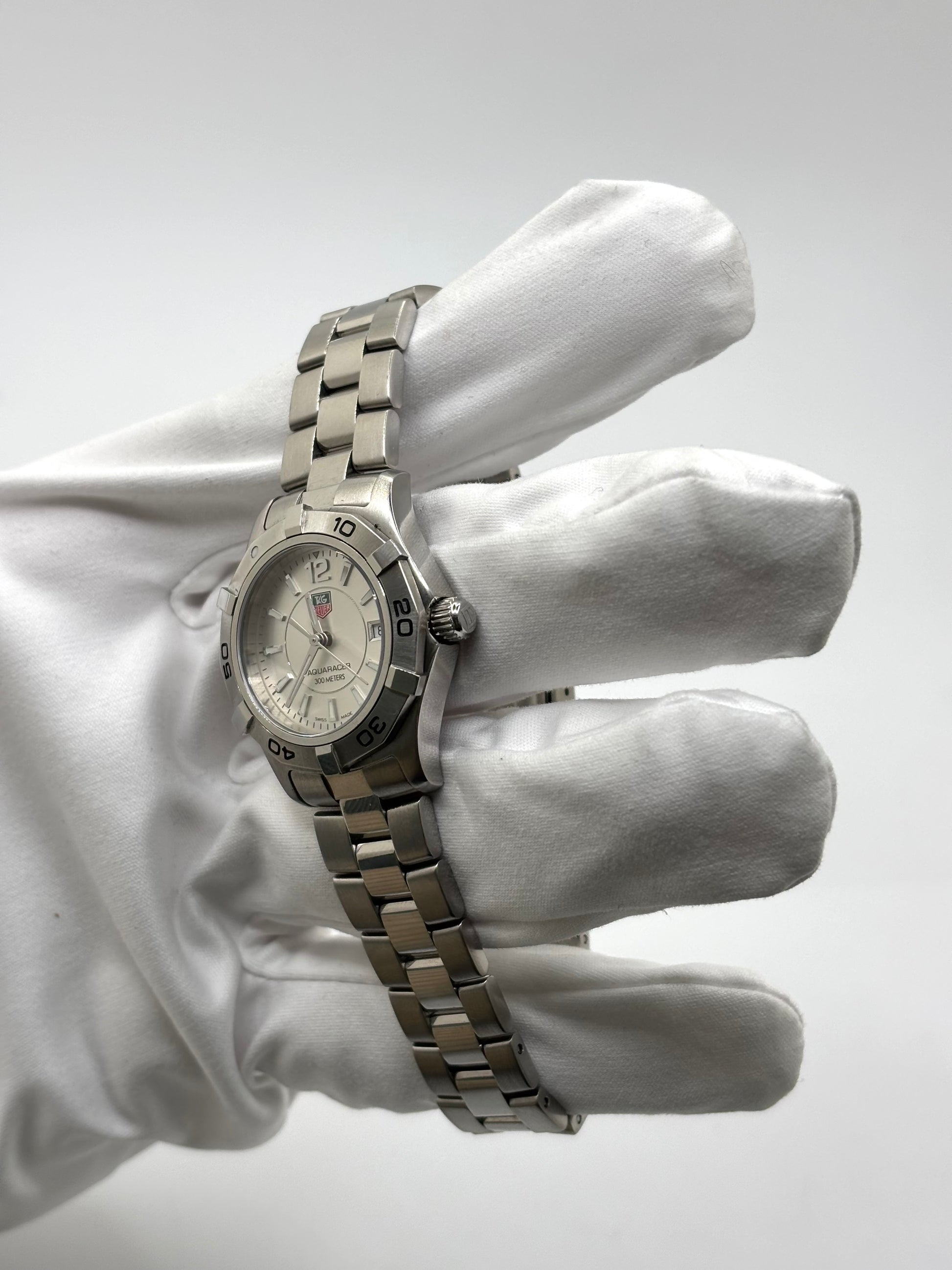 Montre femme TAG Heuer Aquaracer Lady référence WAF1412 en acier avec boîtier 27 mm et bracelet acier. Cadran argent avec chiffres arabes, mouvement quartz précis et étanchéité 30 ATM. Montre de luxe sportive et élégante en très bon état, révisée en 2026, idéale pour un usage quotidien ou une tenue habillée. Détail du boîtier acier poli, montre horlogère suisse féminine, intemporelle et raffinée.