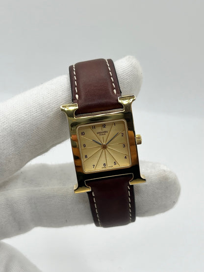 Montre Hermès Heure H HH1.201 pour femme, équipée d’un mouvement quartz et d’un boîtier en acier de 21 mm. Son cadran couleur or est protégé par un verre saphir et accompagné d’un bracelet brun en cuir de vache avec boucle ardillon en acier. Étanche à 3 ATM, cette montre Hermès Heure H se distingue par son célèbre boîtier en forme de H et son design élégant. En très bon état, elle présente de légères traces d’usure.
