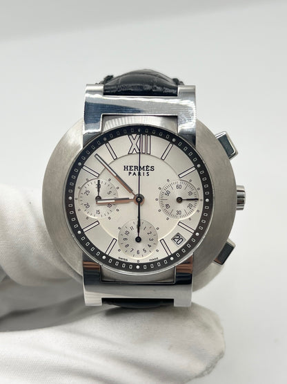 Montre Hermès Nomade Chronograph en acier avec boîtier de 39 mm et cadran blanc à index bâtons. Ce chronographe à mouvement quartz est équipé d’un verre saphir, d’un affichage de la date et d’un bracelet en cuir de crocodile gris avec boucle déployante en acier. Son design élégant et sportif reflète le savoir-faire horloger de la maison Hermès. Modèle unisexe d’occasion en très bon état, alliant confort, précision et raffinement au quotidien.
