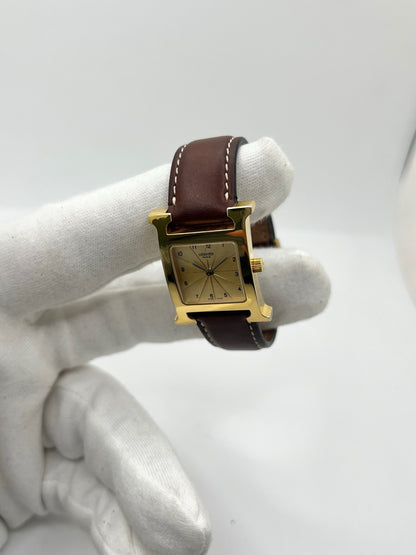 Montre Hermès Heure H HH1.201 pour femme, équipée d’un mouvement quartz et d’un boîtier en acier de 21 mm. Son cadran couleur or est protégé par un verre saphir et accompagné d’un bracelet brun en cuir de vache avec boucle ardillon en acier. Étanche à 3 ATM, cette montre Hermès Heure H se distingue par son célèbre boîtier en forme de H et son design élégant. En très bon état, elle présente de légères traces d’usure.
