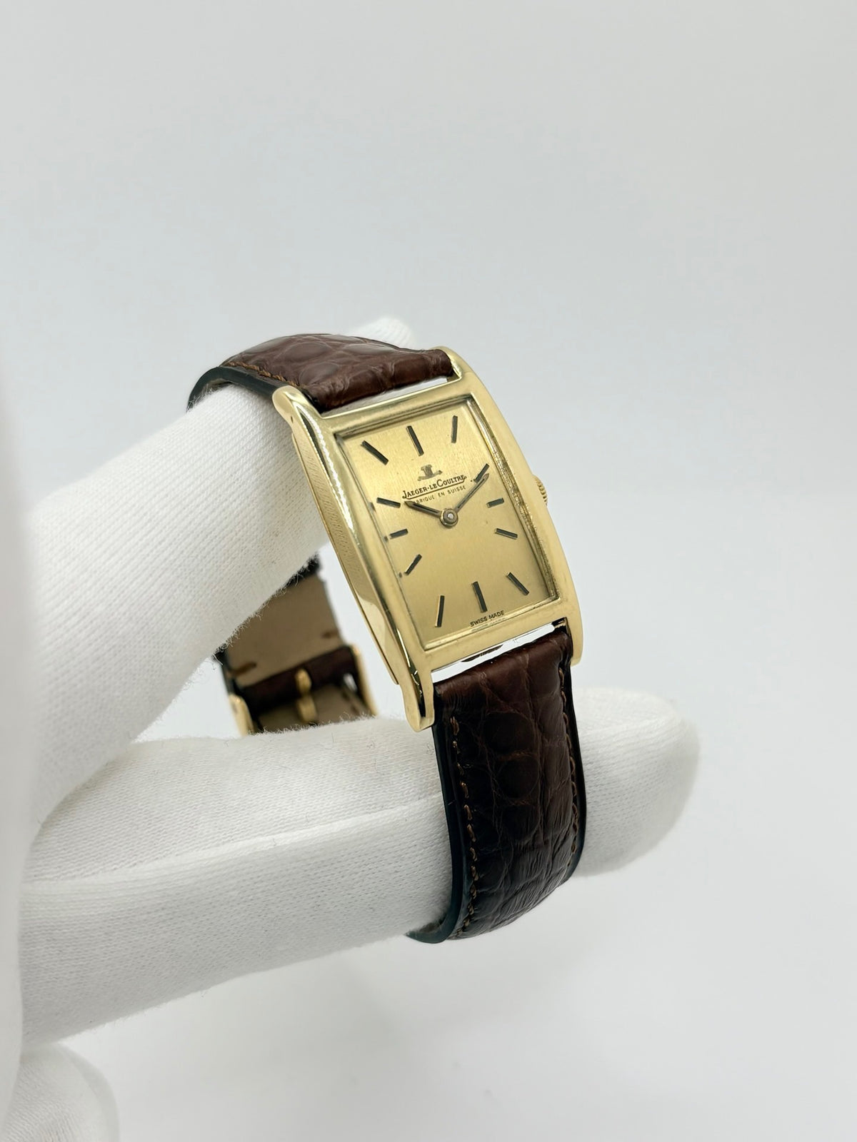 Montre Jaeger-LeCoultre Classic 9012 vintage à remontage manuel, datant approximativement de 1960. Son boîtier rectangulaire en or jaune de 37 × 23 mm est équipé d’un verre Plexiglas et d’un élégant cadran or à index bâtons. Cette montre Jaeger-LeCoultre est montée sur un bracelet brun en cuir de crocodile avec boucle ardillon en acier. En très bon état, elle présente de légères traces d’usure et est proposée avec son coffret d’origine.