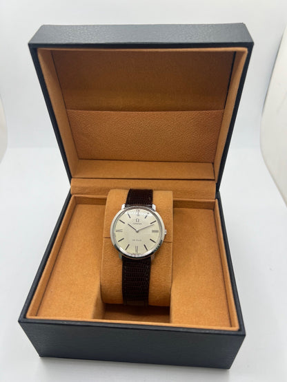 Montre Omega De Ville vintage référence 111.077 en acier, fabriquée en 1969. Ce modèle à remontage manuel est doté d'un cadran argent avec chiffres romains, d'un verre en plexiglas et d'un boîtier de 33 mm. Le bracelet en cuir noir avec boucle ardillon en acier complète son style classique et intemporel. Cette montre d'occasion révisée séduit les amateurs d'horlogerie vintage et de montres Omega de collection.
