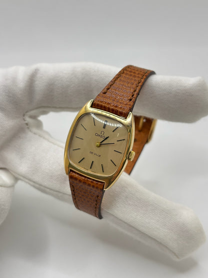 Montre Omega De Ville vintage référence 511.0531 en plaqué or datant de 1969, équipée d'un mouvement à remontage manuel. Son cadran doré à index bâtons est protégé par un verre en matière plastique. Son boîtier de 25 mm est associé à un bracelet en cuir brun avec boucle ardillon en acier. Cette montre d'occasion, révisée et en très bon état, séduit par son élégance intemporelle et son style classique des années 1960.