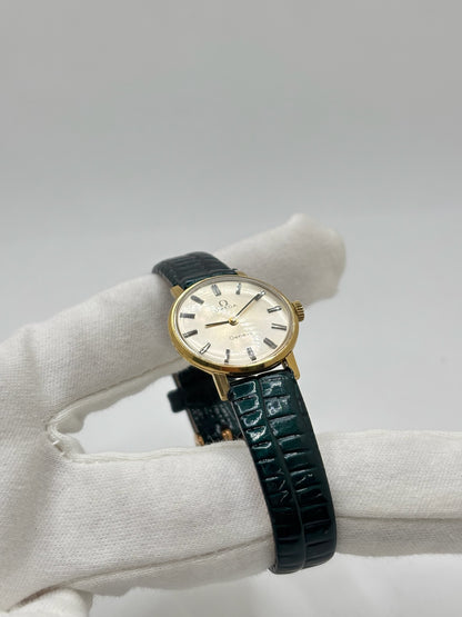 Montre Omega Genève vintage référence 511.0457 pour femme, fabriquée vers 1972 et équipée d'un mouvement à remontage manuel. Son boîtier en acier de 23 mm est associé à un cadran blanc à index bâtons protégé par un verre en matière plastique. Elle est montée sur un bracelet en cuir vert avec boucle ardillon en acier. Révisée en interne et proposée en très bon état, cette montre d'occasion incarne l'élégance discrète et le savoir-faire horloger suisse des années 1970.