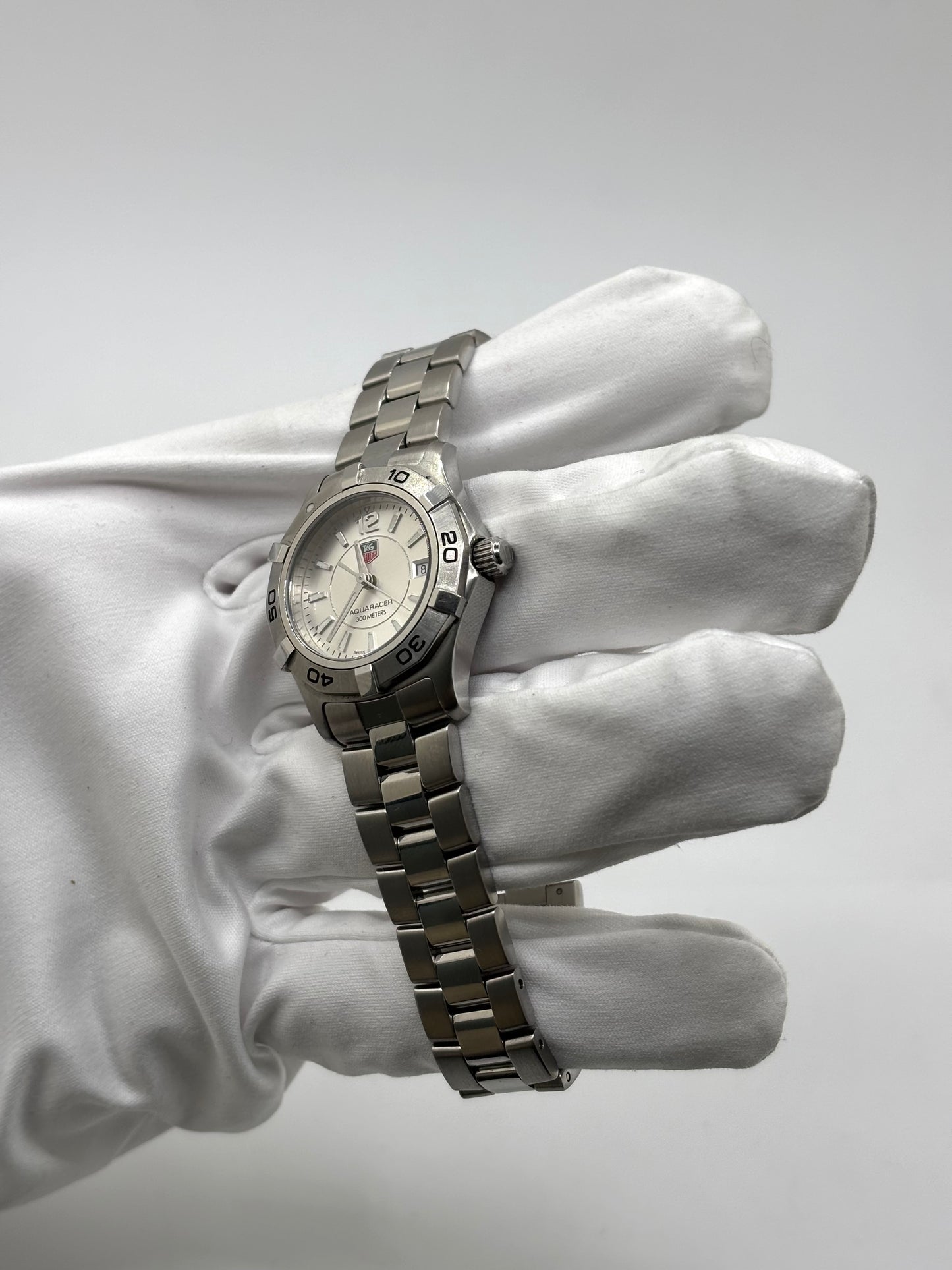 Montre femme TAG Heuer Aquaracer Lady référence WAF1412 en acier avec boîtier 27 mm et bracelet acier. Cadran argent avec chiffres arabes, mouvement quartz précis et étanchéité 30 ATM. Montre de luxe sportive et élégante en très bon état, révisée en 2026, idéale pour un usage quotidien ou une tenue habillée. Détail du boîtier acier poli, montre horlogère suisse féminine, intemporelle et raffinée.