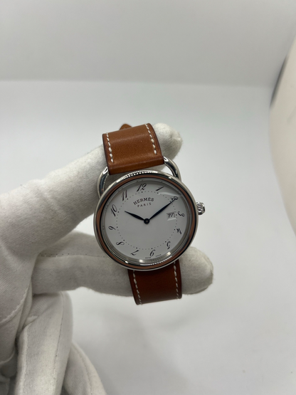 Montre Hermès Arceau Grand Modèle AR5.710 de 2014 en acier 38 mm avec mouvement quartz, cadran blanc à chiffres arabes et bracelet cuir brun d’origine. Cette montre de luxe Hermès Full Set avec boîte et papiers possède un verre saphir, une boucle ardillon acier et le design emblématique Arceau inspiré de l’univers équestre. Montre vintage Hermès élégante, révisée en 2026 et garantie 1 an.