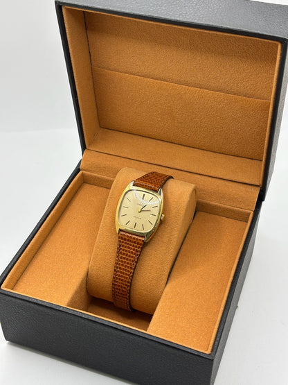 Montre Omega De Ville vintage référence 511.0531 en plaqué or datant de 1969, équipée d'un mouvement à remontage manuel. Son cadran doré à index bâtons est protégé par un verre en matière plastique. Son boîtier de 25 mm est associé à un bracelet en cuir brun avec boucle ardillon en acier. Cette montre d'occasion, révisée et en très bon état, séduit par son élégance intemporelle et son style classique des années 1960.