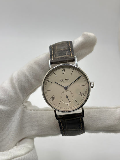 Montre Nomos Glashütte Ludwig 205 en acier avec boîtier de 37,5 mm, mouvement à remontage manuel offrant 43 heures de réserve de marche et cadran blanc à chiffres romains. Équipée d'un verre saphir, d'un bracelet en cuir noir, d'une petite seconde, d'un fond transparent et d'aiguilles en acier bleui, cette montre de luxe d'occasion est en très bon état avec de légères traces d'usure. Un modèle élégant au style intemporel, idéal pour les amateurs d'horlogerie.
