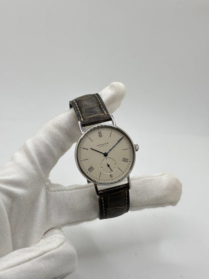 Montre Nomos Glashütte Ludwig 205 en acier avec boîtier de 37,5 mm, mouvement à remontage manuel offrant 43 heures de réserve de marche et cadran blanc à chiffres romains. Équipée d'un verre saphir, d'un bracelet en cuir noir, d'une petite seconde, d'un fond transparent et d'aiguilles en acier bleui, cette montre de luxe d'occasion est en très bon état avec de légères traces d'usure. Un modèle élégant au style intemporel, idéal pour les amateurs d'horlogerie.
