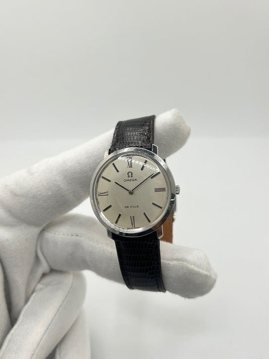 Montre Omega De Ville vintage référence 111.077 en acier, fabriquée en 1969. Ce modèle à remontage manuel est doté d'un cadran argent avec chiffres romains, d'un verre en plexiglas et d'un boîtier de 33 mm. Le bracelet en cuir noir avec boucle ardillon en acier complète son style classique et intemporel. Cette montre d'occasion révisée séduit les amateurs d'horlogerie vintage et de montres Omega de collection.
