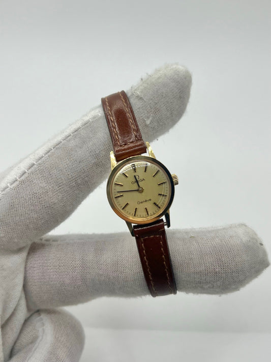 Montre Omega Genève Lady en acier avec boîtier plaqué or de 20 mm, cadran or à index bâtons, mouvement calibre 560, bracelet en cuir brun avec boucle acier. Vintage de 1978, d'occasion en bon état avec traces d'usure visibles. Verre plexiglas, modèle élégant pour femme, idéal pour collection ou usage quotidien. Livrée sans coffret ni papiers d'origine, disponible en stock à Paris.