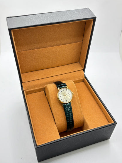 Montre Omega Genève vintage référence 511.0457 pour femme, fabriquée vers 1972 et équipée d'un mouvement à remontage manuel. Son boîtier en acier de 23 mm est associé à un cadran blanc à index bâtons protégé par un verre en matière plastique. Elle est montée sur un bracelet en cuir vert avec boucle ardillon en acier. Révisée en interne et proposée en très bon état, cette montre d'occasion incarne l'élégance discrète et le savoir-faire horloger suisse des années 1970.