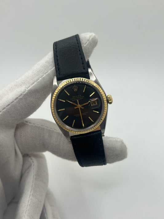 Montre Rolex Datejust automatique vintage de 36 mm en acier avec lunette en or jaune, cadran noir épuré sans chiffres et affichage de la date à 3 heures. Boîtier élégant protégé par un verre plexiglas, étanche jusqu’à 10 ATM. Bracelet en cuir noir avec boucle en acier. Mouvement mécanique à remontage automatique offrant environ 40 heures de réserve de marche. Modèle des années 1960, en très bon état, présentant de légères traces d’usure superficielles.