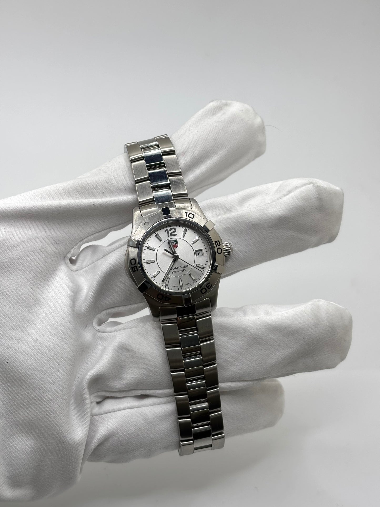 Montre femme TAG Heuer Aquaracer Lady référence WAF1412 en acier avec boîtier 27 mm et bracelet acier. Cadran argent avec chiffres arabes, mouvement quartz précis et étanchéité 30 ATM. Montre de luxe sportive et élégante en très bon état, révisée en 2026, idéale pour un usage quotidien ou une tenue habillée. Détail du boîtier acier poli, montre horlogère suisse féminine, intemporelle et raffinée.